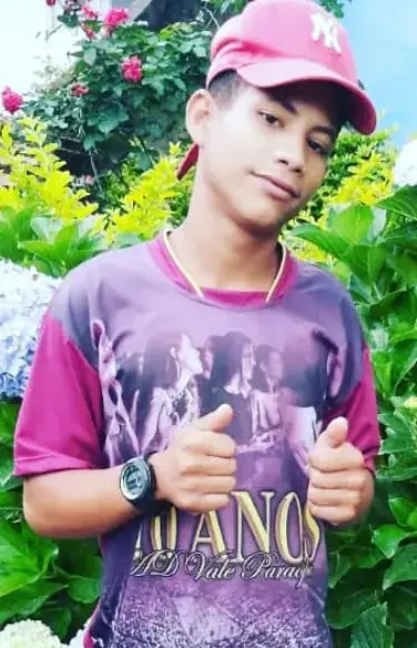 Imagem de um jovem (Frederico Gonçalves dos Santos), com as duas mãos fazendo sinal de positivo e no fundo um jardim florido.