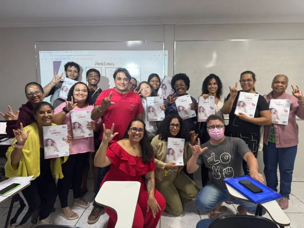Cerca de 16 pessoas em uma sala de aula da turma do curso de Libras, fazendo o sinal com a mão que significa AMOR.
