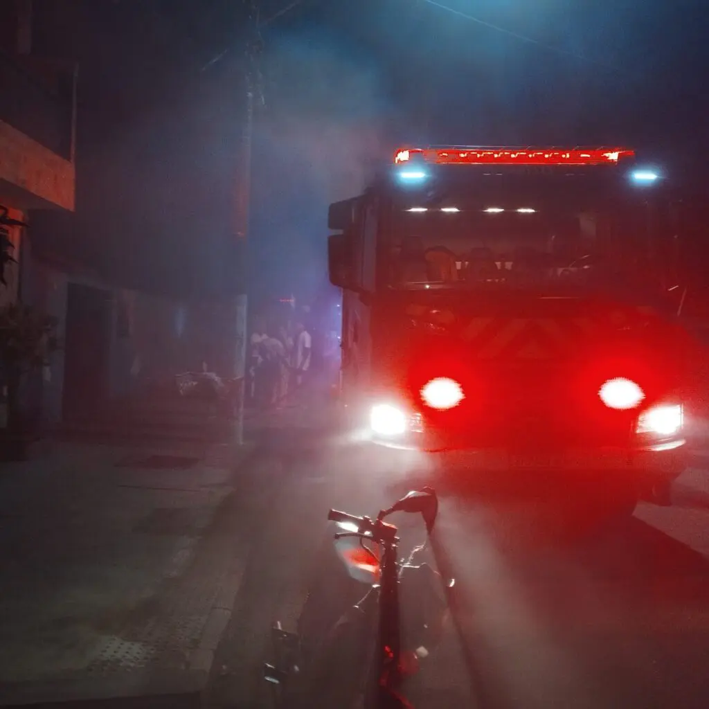 Um caminhão do Corpo de Bombeiros parado em uma rua residencial durante a noite em volto em fumaça vinda de uma casa que estava em chamas.