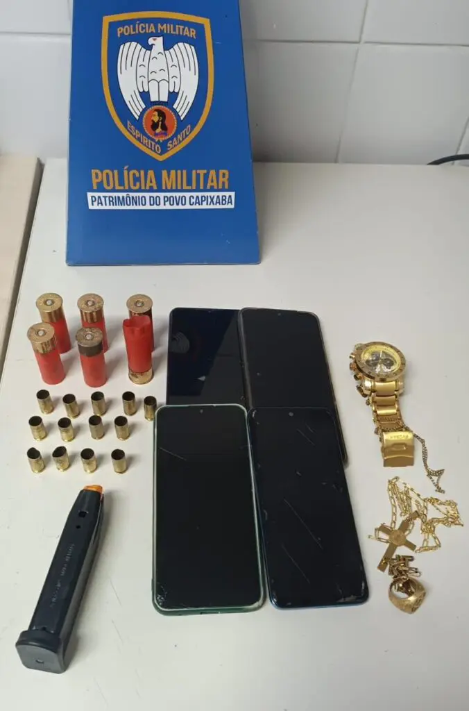 Imagem de uma mesa com alguns itens apreendidos pela Polícia Militar, sobre a mesa há quatro celulares, um relógio dourado, uma corrente dourada, um carregador de pistola, duas balas calibre 12 intactas, três mastigadas, e uma deflagrada e 13 capsulas de bala calibre .40.