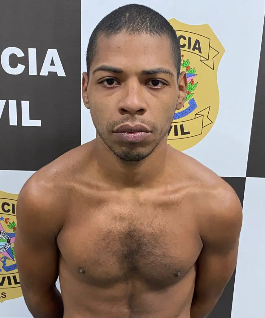 Imagem de um homem com os braços para trás parado em frente a um banner da Polícia Civil.
