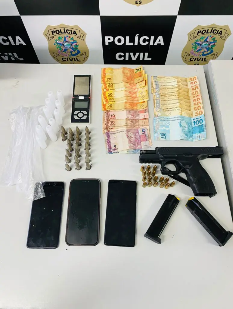 Imagem de materiais apreendidos pela PC, sobre uma mesa três celulares, uma pistola, dois carregadores, 17 munições, cerca de 957 reais em espécie, 23 pinos de cocaína, uma balança de precisão além de algum materiais para embalo de drogas.