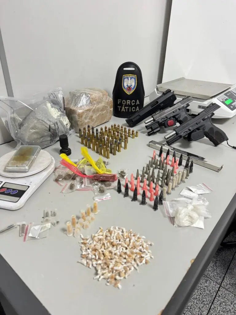 Imagem de vários materiais apreendidos pela Polícia Militar sobre uma mesa, duas pistolas, um simulacro de pistola, munições e diversos porções de drogas.