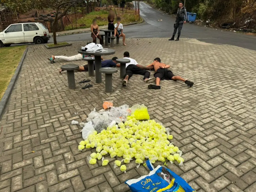 Em uma praça um policial militar, parado ao lado de três mulheres detidas, quatro homens deitados no chão e 6.721 pinos de cocaína espalhados durante uma ação da Polícia Militar.