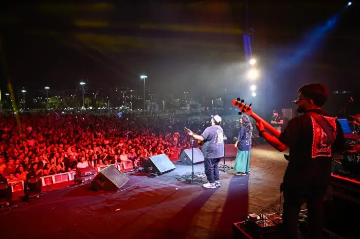 Imagem de um palco visto dos bastidores com os músicos de uma banda se apresentando para uma plateia lotada.