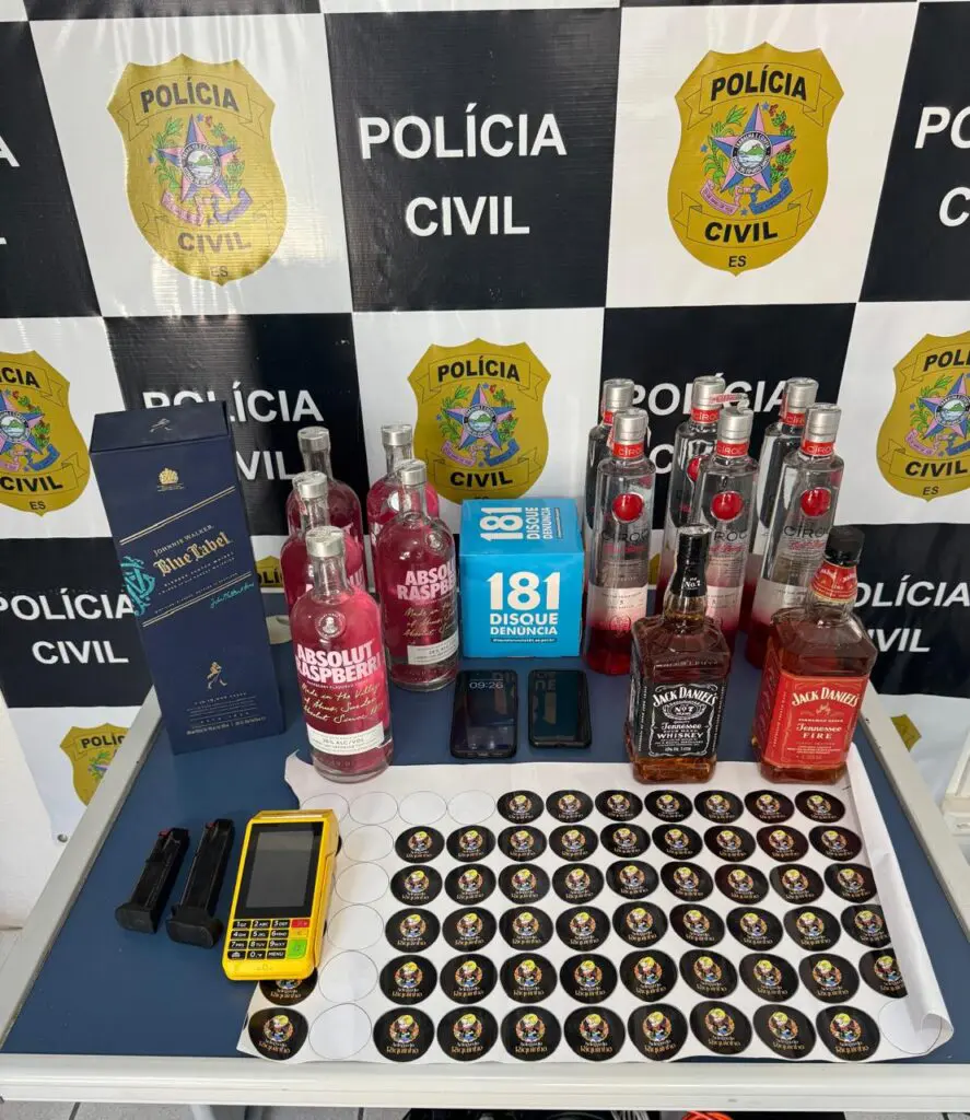 Imagem de uma mesa com diversas garrafas de bebidas alcoólicas falsificadas, dois celulares, uma máquina de cartões, dois carregadores de arma, dois celulares e adesivos falsos.