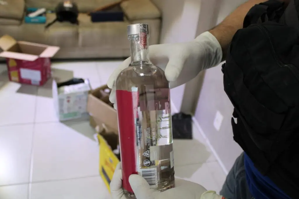 Em uma sala com diversas caixas de bebidas alcoólicas, um policial civil segurando uma garrafa de bebida alcoólica falsificada.