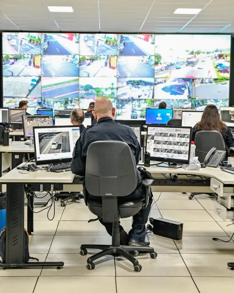 Imagem de uma sala de videomonitoramento com um telão exibindo diversas câmeras de segurança espalhadas pela cidade, e cerca de seis funcionários posicionados em frente a monitores de videomonitoramento.