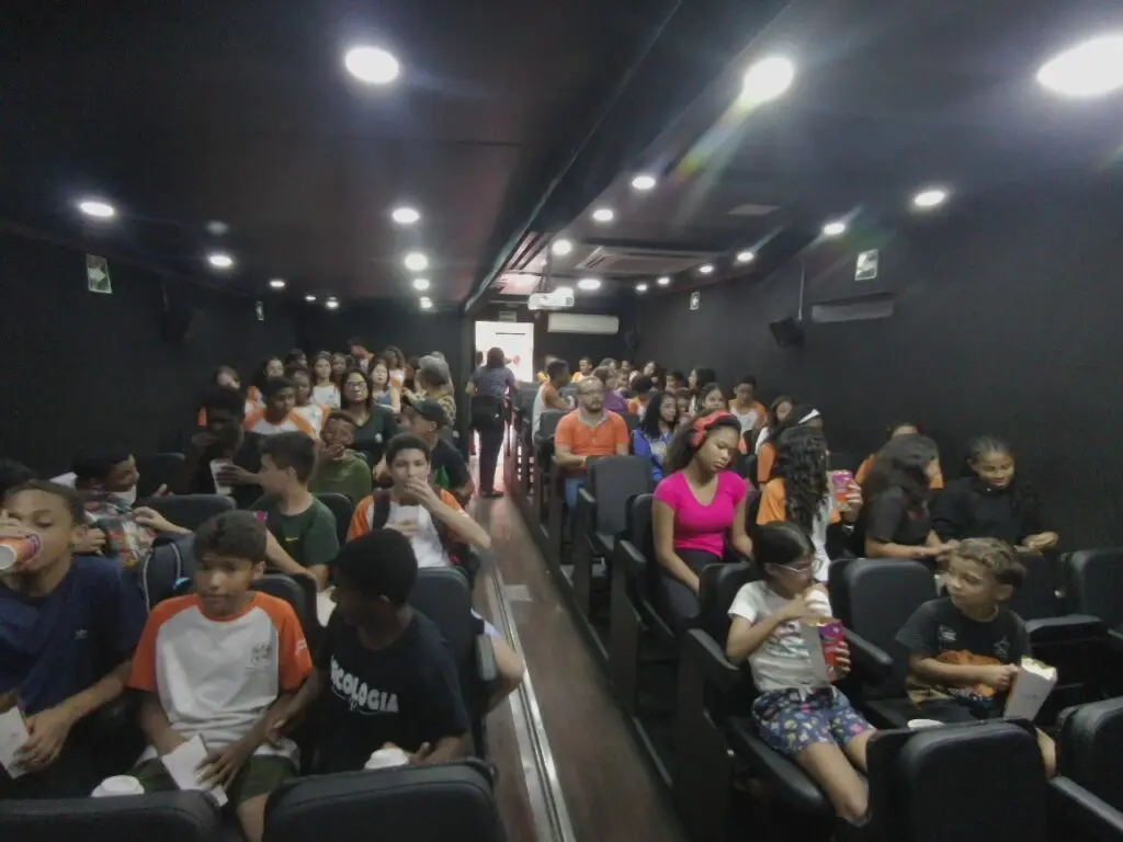 Imagem de uma sala de cinema dentro de uma carreta, com diversas pessoas acomodadas em suas poltronas. 