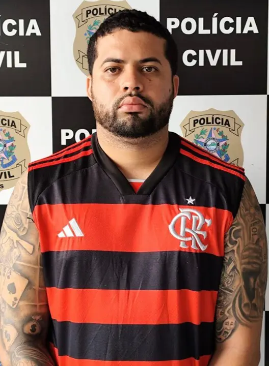 Imagem de um homem (Felipe Barreto de Assis, conhecido como "Barretinho", de 27 anos) em frente a uma parede com imagens de divulgação da Polícia Civil.