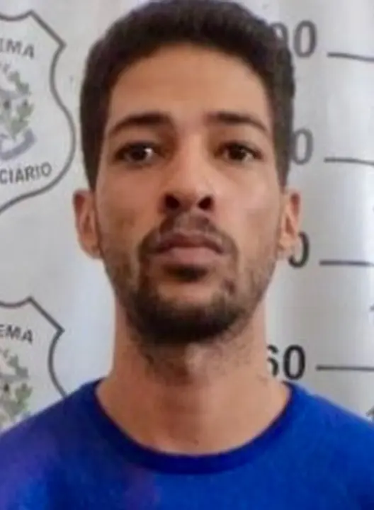 Imagem de um homem (Luiz Henrique Batista da Silva) em frente a uma parede com imagens de divulgação da Polícia Civil.