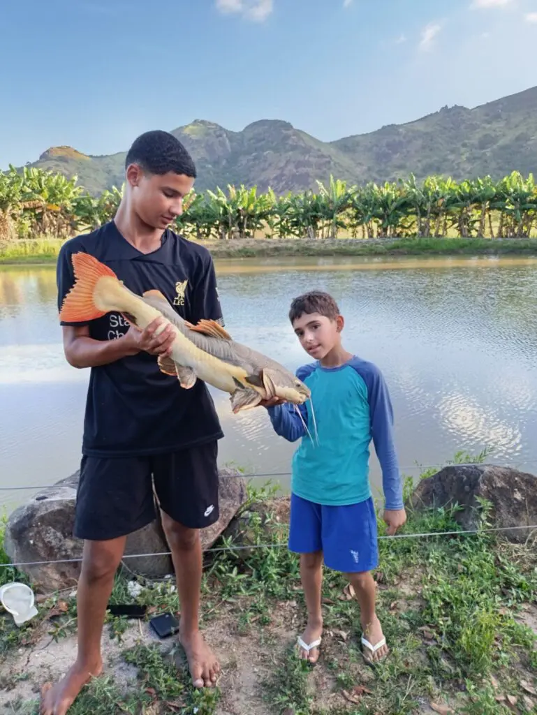 Imagem de um adolescente segurando um peixe da espécie Pirarara ao lado de uma criança, no fundo um lago.