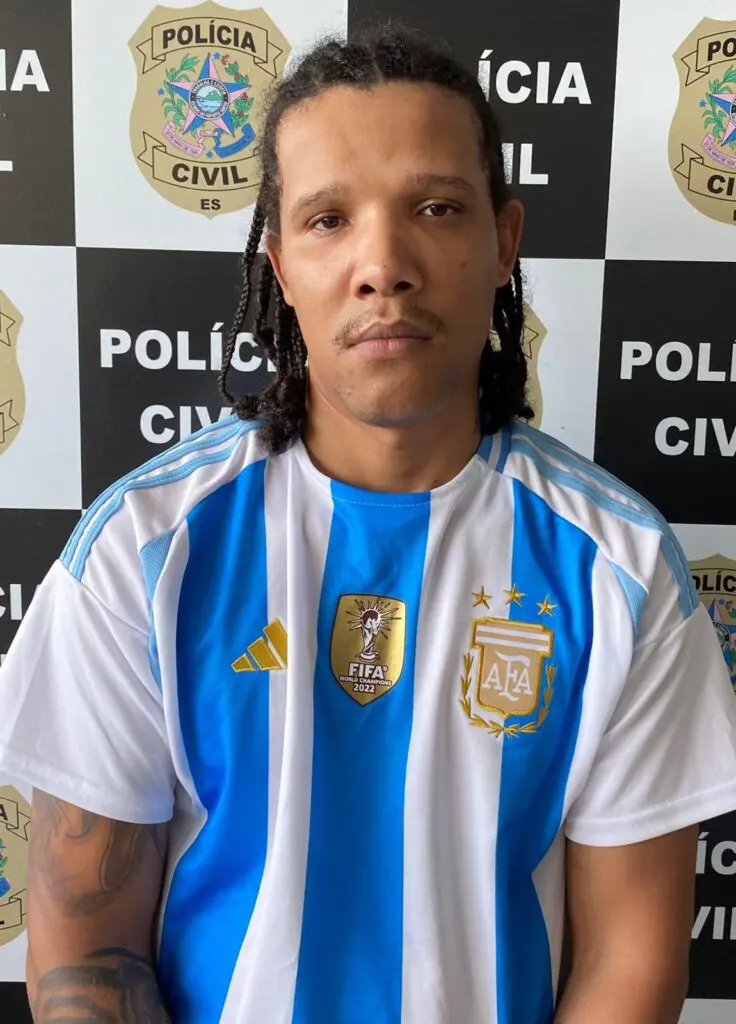 Imagem de um homem (Lázaro Silva Muniz, vulgo “Popeye”, 28 anos) em frente a uma parede com imagens de divulgação da Polícia Civil.