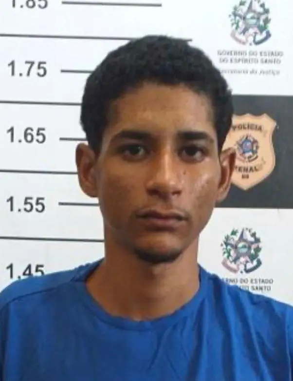 Imagem de um homem (Luiz Gustavo de Souza Sobrinho, vulgo 
“Gustavinho”, 21 anos) em frente a uma parede com imagens de divulgação da Polícia Civil.