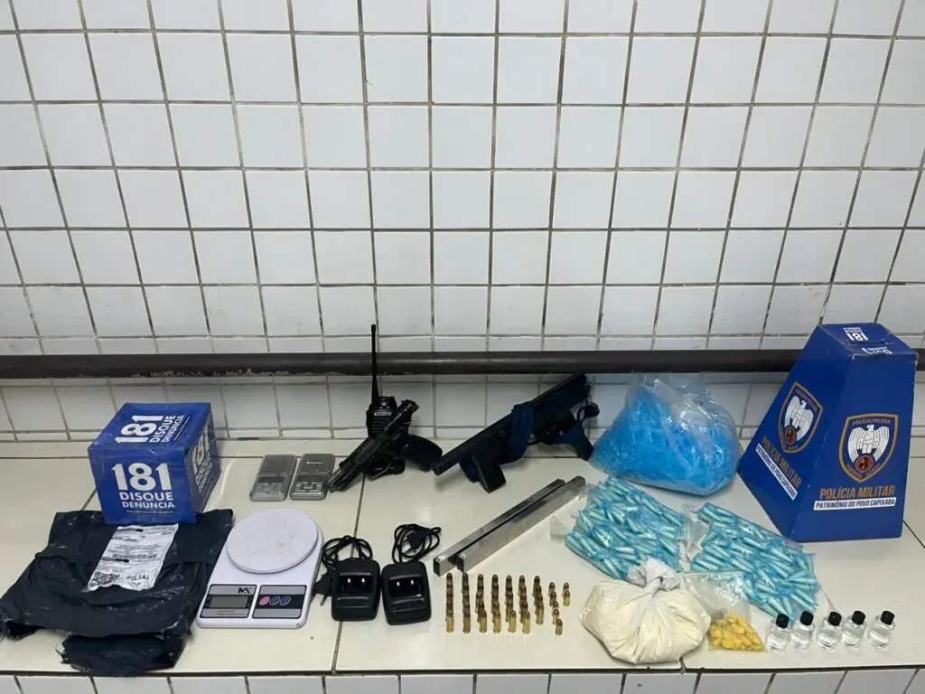 Sobre uma bancada materiais apreendidos pela PM: 286 pinos de cocaína, uma porção maior da droga pesando 426 gramas, 29 comprimidos de ecstasy, cinco frascos de loló, um rádio comunicador com base, uma pistola calibre 9mm, uma metralhadora 9mm com dois carregadores, 37 munições calibre 38, sete munições calibre 380.