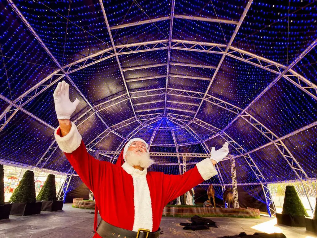 Imagem de um homem fantasiado de Papai Noel de braços abertos olhando para cima em um local totalmente decorado com leds.