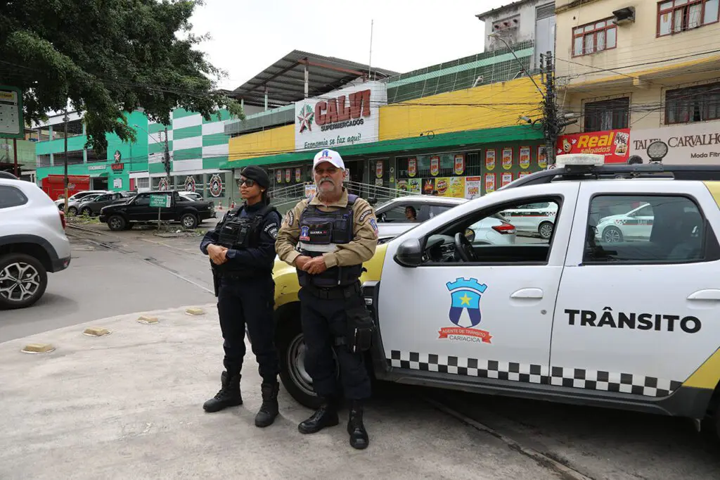 Imagem de dois agentes de trânsito próximos de uma viatura em um local com diversos comércios. 