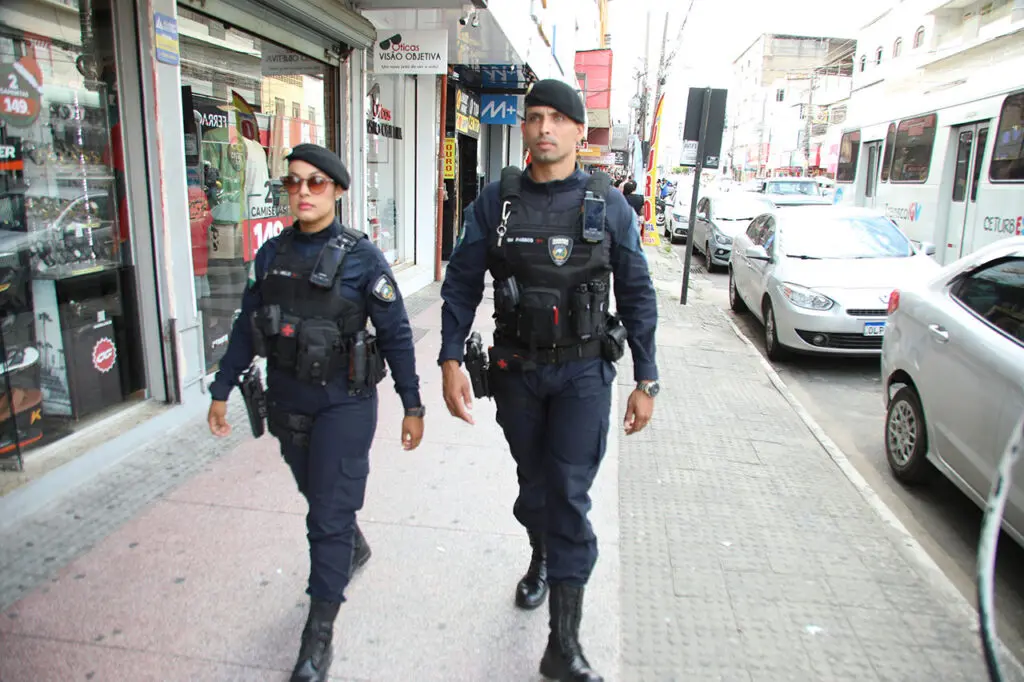 Imagem de dois agentes da Guarda de Cariacica andando em uma rua com diversos comércios.