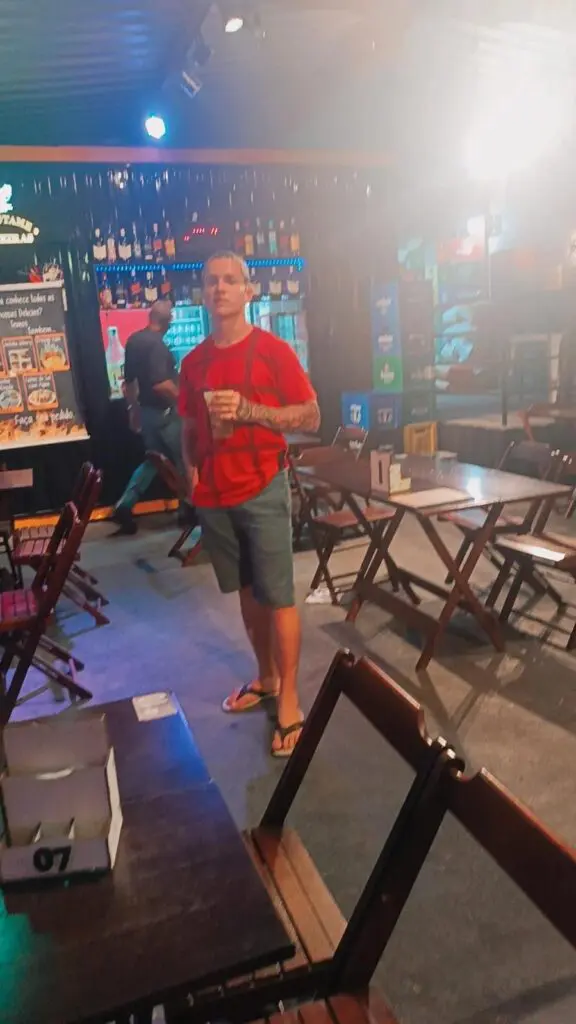 Um jovem (Gilvan Rodrigues de Oliveira Junior) segurando um copo com bebida, em um bar.