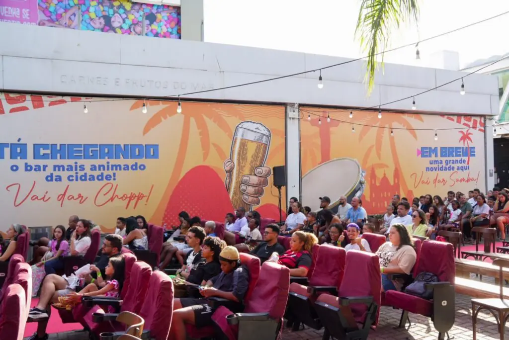 Imagem de um telão de cinema instalado em um local a ar livre, com diversas pessoas assistindo a um filme.