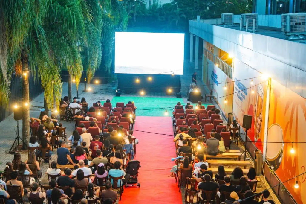 Imagem de um telão de cinema instalado em um local a ar livre, com diversas pessoas assistindo a um filme.