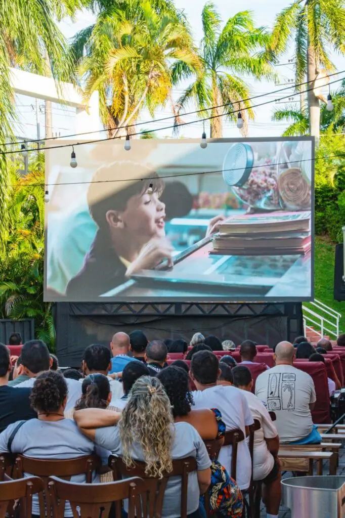 Imagem de um telão de cinema instalado em um local a ar livre, com diversas pessoas assistindo a um filme.
