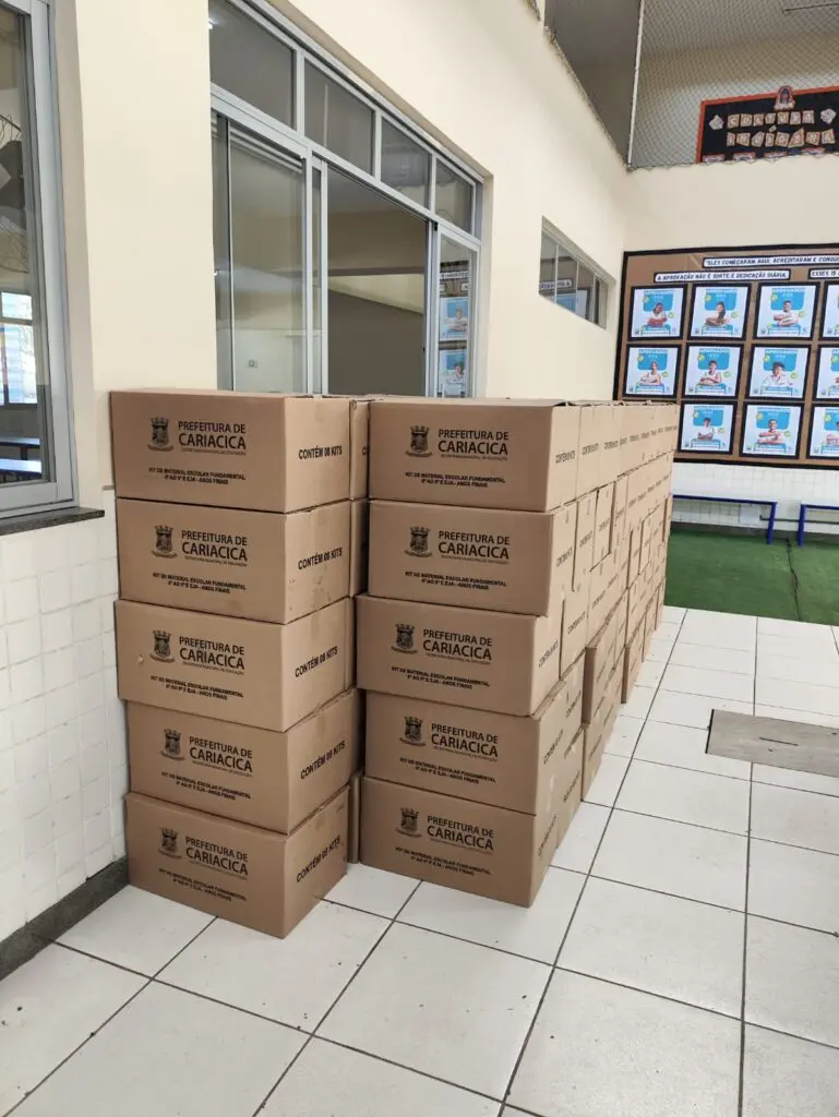 Um corredor de uma escola com duas fileiras de caixas de materiais escolares com cerca de 100 caixas, sobre o chão.