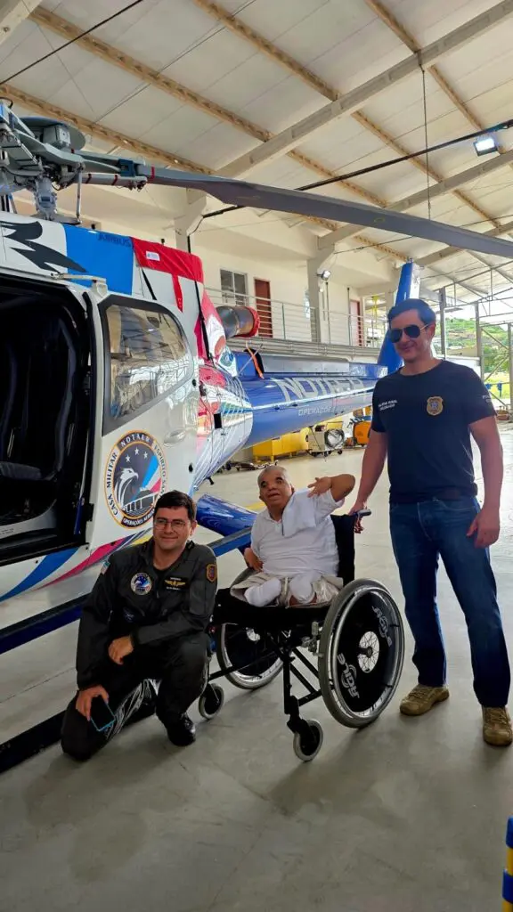 Foto de dois delegados ao lado de um jovem em uma cadeira de rodas, fazendo um sinal de positivo, posando para uma foto ao lado de um helicóptero.