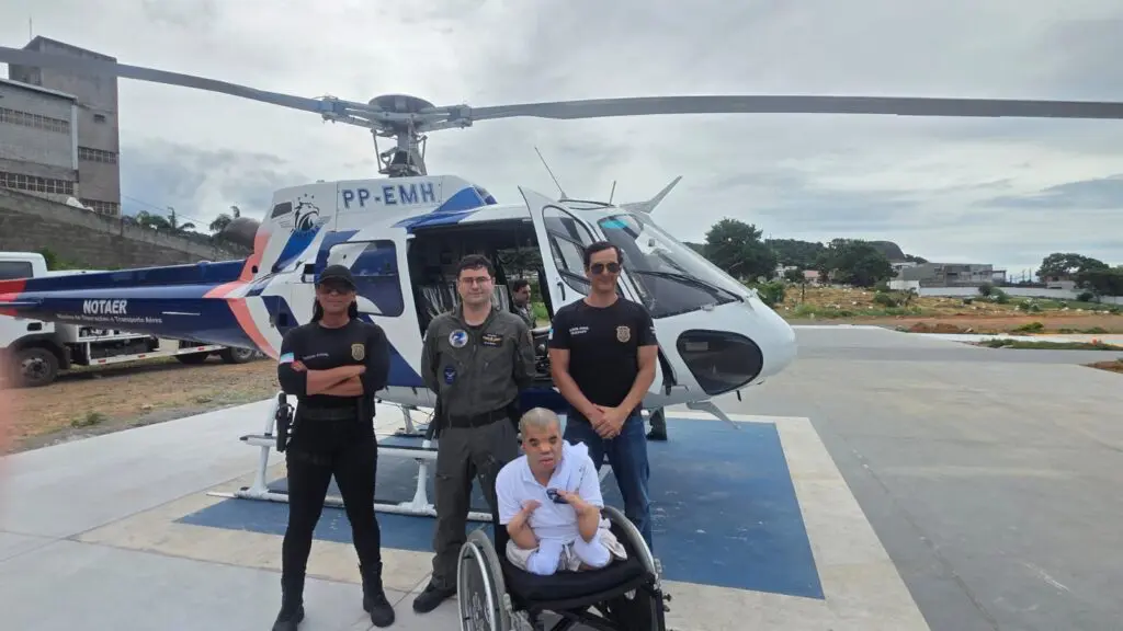 Foto de dois policiais civis ao lado de um piloto e de um jovem em uma cadeira de rodas, fazendo um sinal de positivo, posando para uma foto ao lado de um helicóptero.