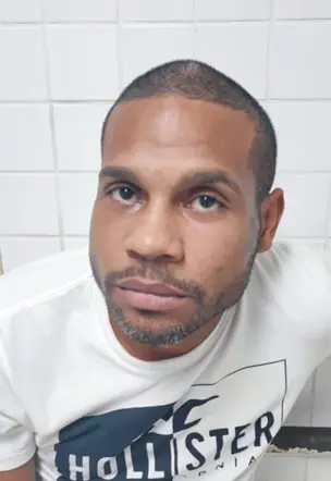 Imagem de um sujeito (Carlos Alberto dos Santos Gonçalves Júnior, conhecido como “Bequinha”), algemado com as mãos para trás.