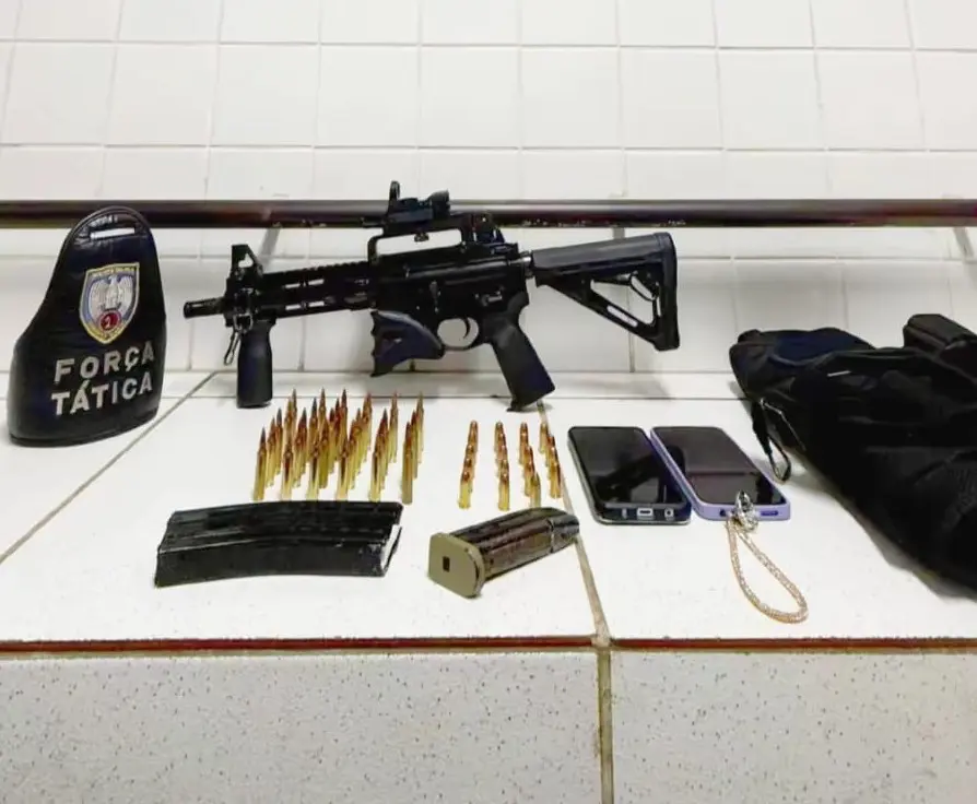 Imagem de materiais apreendidos pela PM sobre uma bancada: um fuzil calibre 5.56, dois carregadores, munições, dois celulares e uma capa de colete balistico.