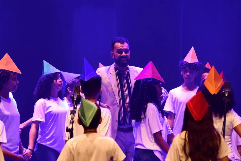 Imagem de um professor e em volta, diversas crianças com chapéus feitos de papel.