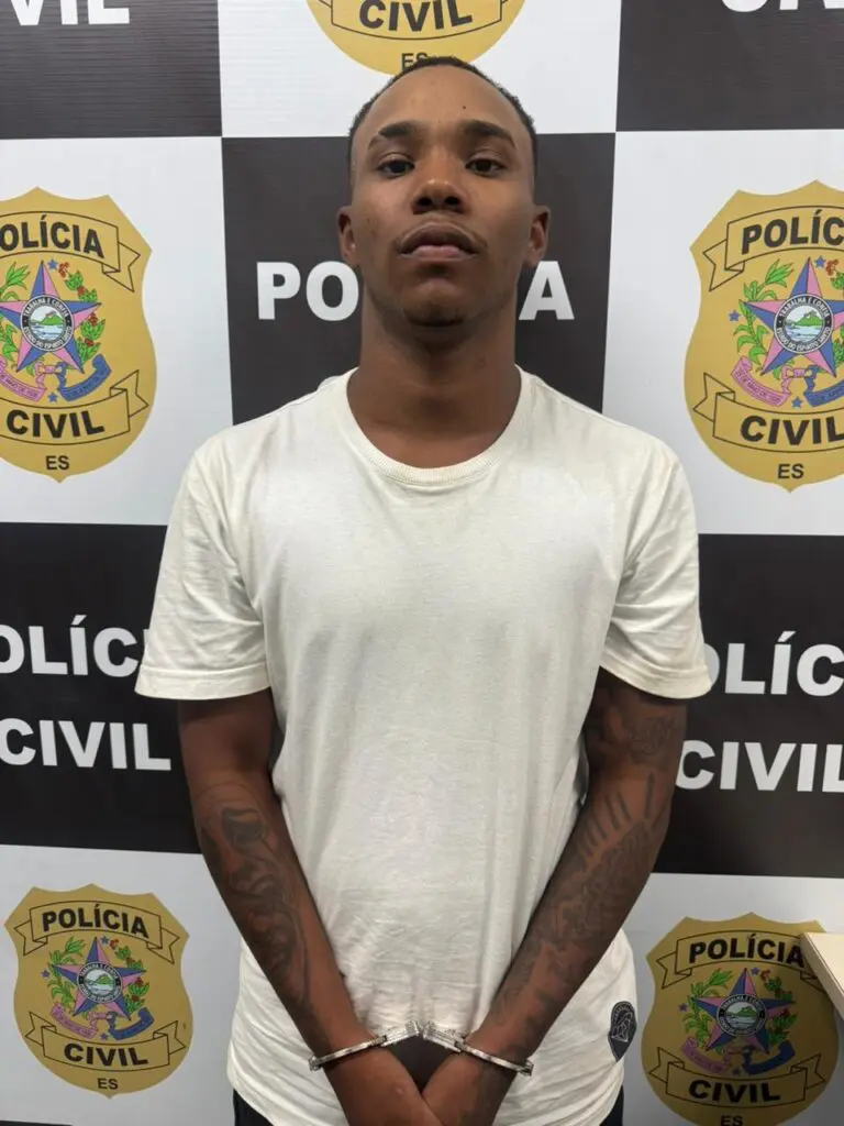 Imagem de um jovem (Kauã Vital dos Santos, de 18 anos) com as mãos para frente, algemado em frente a uma parede com identificações da Polícia Civil.
