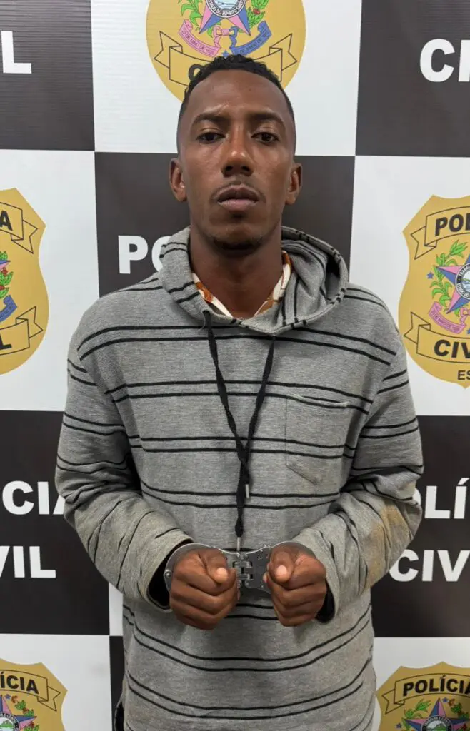 Imagem de um homem (Eric Gordiano dos Santos, de 26 anos) com as mãos para frente, algemado em frente a uma parede com identificações da Polícia Civil.