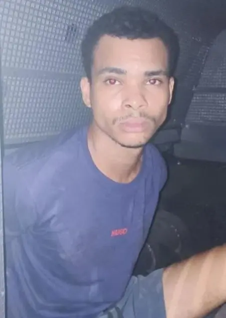Imagem de um sujeito (Leonardo Julio Lopes, de 23 anos), preso pelo crime de latrocínio, sentado algemado com as mãos para trás, dentro de um compartimento de segurança de uma viatura da polícia.