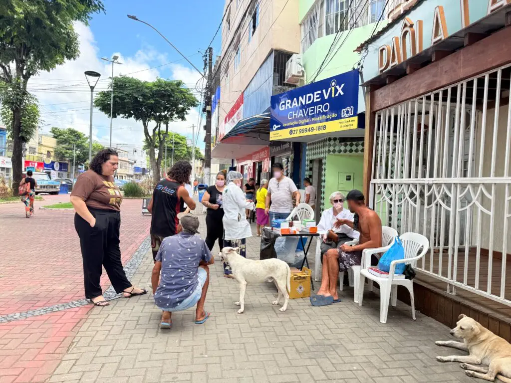 Imagem de uma praça, com assistentes sociais e uma equipe médica realizando exames em pessoas em condição de rua.