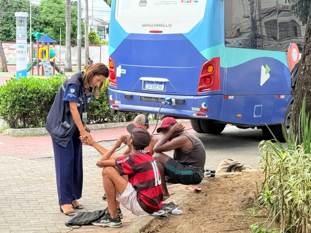 Imagem de uma praça, com uma assistente social apertando a mão de um homem em situação de rua.