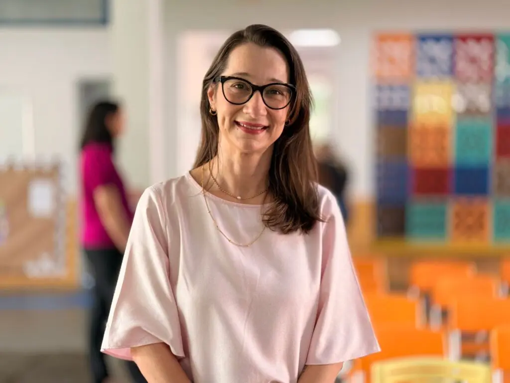 Imagem de uma mulher usando óculos, sorrindo, e no fundo desfocada uma sala de aula.