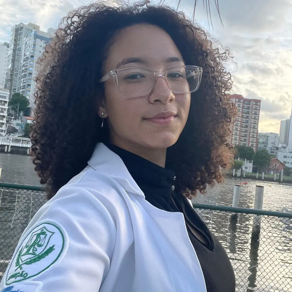 Imagem de uma jovem (Cynthia Beatriz Vasconcellos da Silva Fagundes, de 20 anos) de jaleco, posando para uma foto, e no fundo, uma lagoa e prédios residenciais.