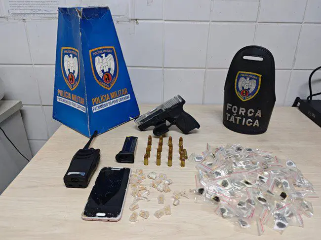 Sobre uma mesa, uma pistola calibre 9mm, um carregador sobressalente, munições de diferentes calibres, incluindo .380 e .38, um aparelho celular, um rádio comunicador, 26 pedras de crack e 93 unidades de haxixe.