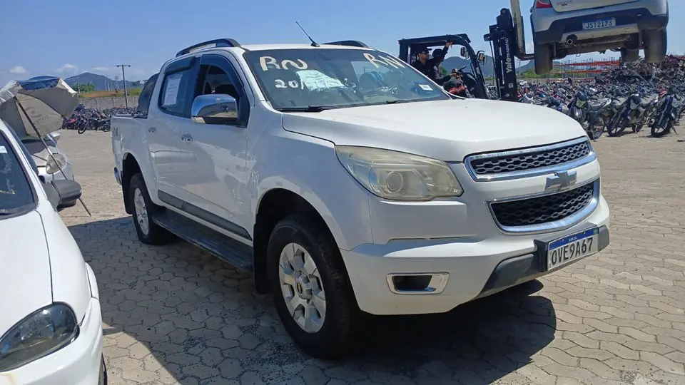 Picape Chevrolet S10, ano 2013, estacionada em um pátio, ao lado de outros veículos.