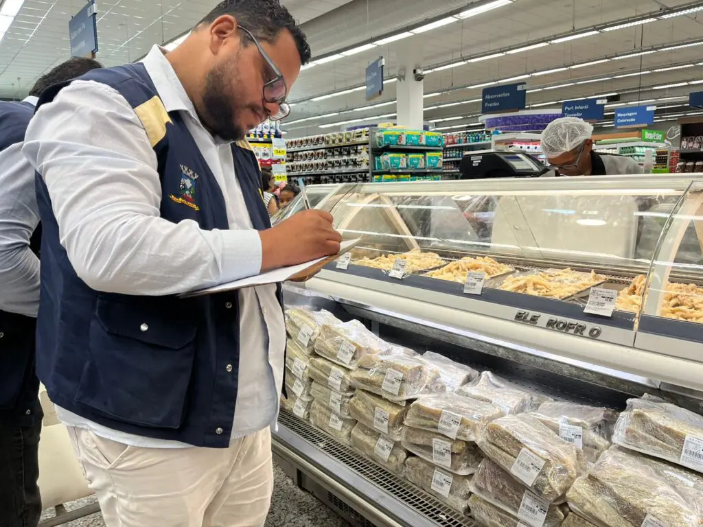 Um fiscal do Procon de Cariacica segurando uma prancheta em frente a um balcão de exposição de bacalhau.