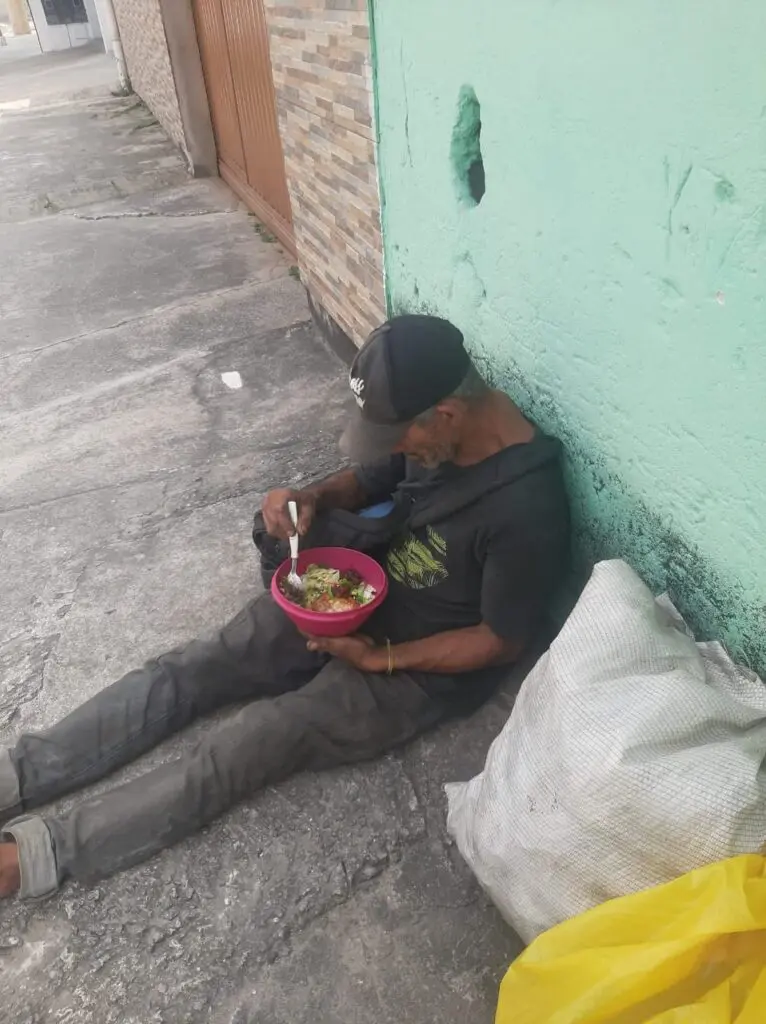 Um homem sentado em uma calçada encostado em um muro, segurando uma bacia com comida.