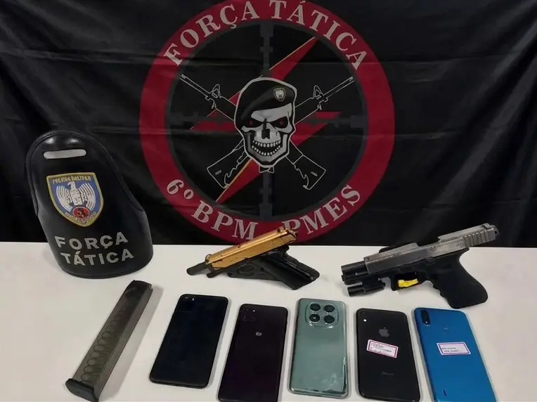 Sobre uma mesa, duas pistolas, cinco aparelhos celulares e um carregador alongado. E na parede, a bandeira da Força Tática do 6º Batalhão, com uma caveira de boina e dois fuzis atravessados em X atrás da caveira.