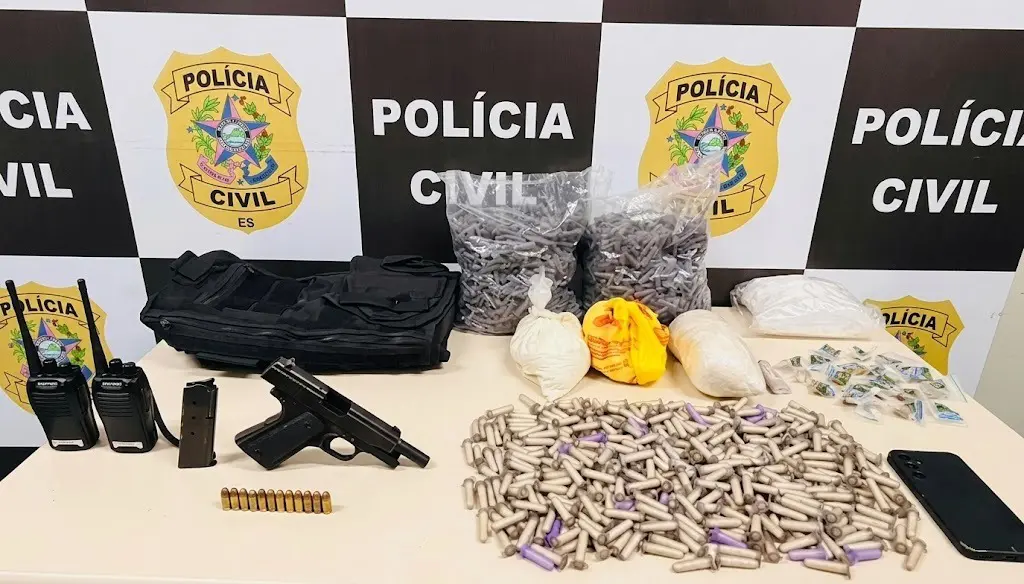 Sobre uma mesa, uma pistola, dois rádios comunicadores, pinos de cocaína, materiais utilizados para embalar drogas, um celular e um artefato explosivo.
