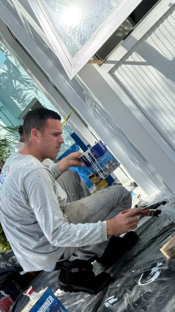 Foto de um homem segurando um pincel e uma lata de tinta, próximo a uma parede.