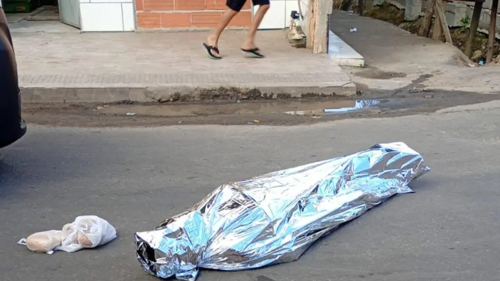 Corpo de um homem coberto caido na pista ao lado de uma sacola com pães.