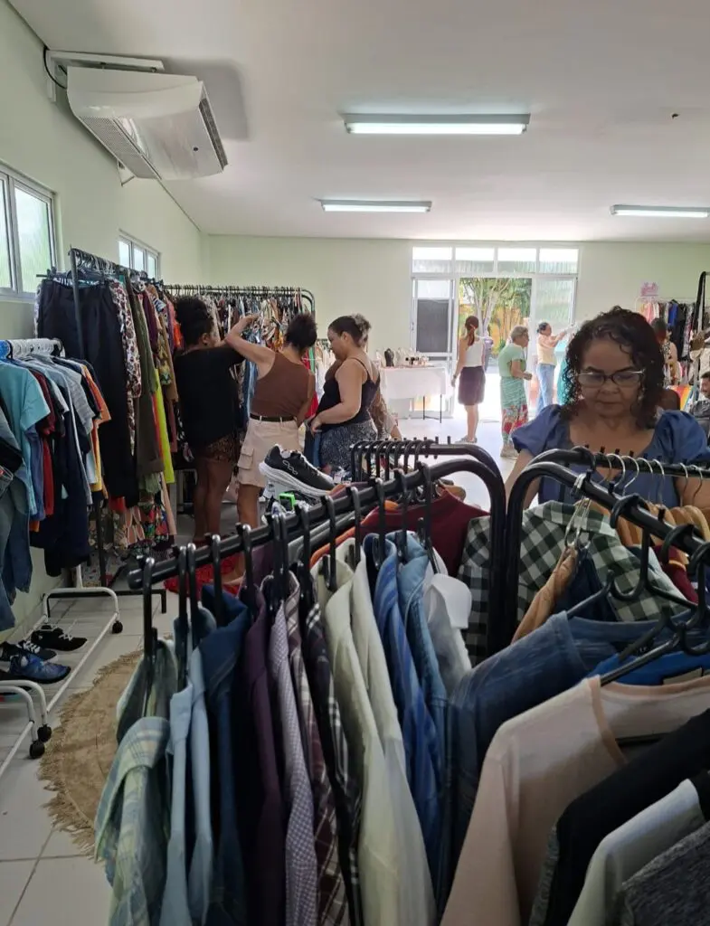 Foto de uma das salas utilizadas pelas expositoras, onde está montada uma loja com várias araras de roupas expostas em um brechó, e cerca de seis mulheres escolhendo algumas peças de roupa.
