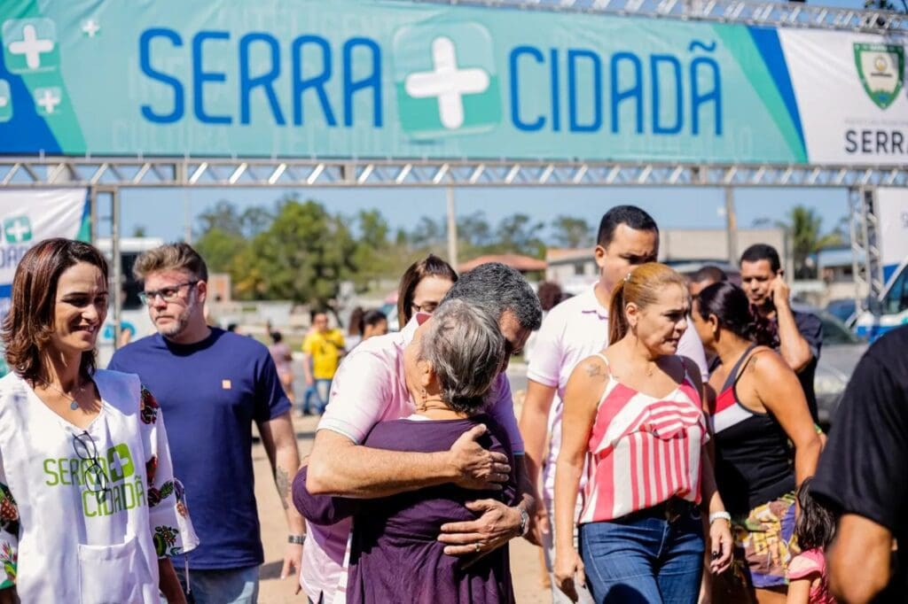 Prefeito Sérgio Vidigal abraçando uma senhora em frente a placa de divulgação do Serra mais Cidadã