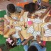 Foto ilustrativa de crianças em volta de uma mesa realizando pinturas em papel, com lápis coloridos